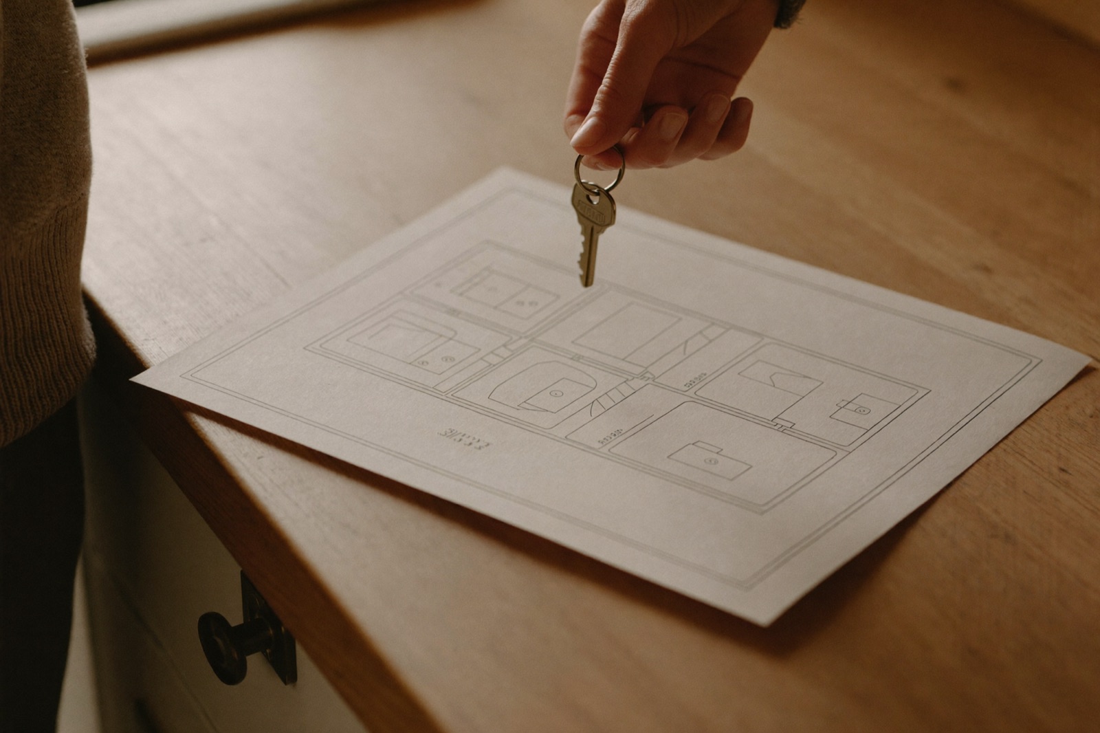 A hand holding a small ring of brass house keys above a kitchen worktop with a folded floor plan.
